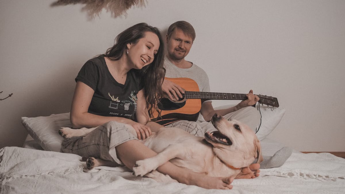 A couple enjoys a cozy moment playing guitar and relaxing with their dog on a bed indoors.